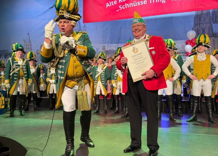 ChatGPT: Das Foto zeigt eine Ehrung bei der Alten Kölner Karnevalsgesellschaft „Schnüsse Tring“ 1901 e.V.. Ein Mann in roter Jacke erhält eine Urkunde, während neben ihm ein Gardist in grüner Uniform mit Federhut spricht. Im Hintergrund stehen zahlreiche Mitglieder der Gesellschaft auf der Bühne.