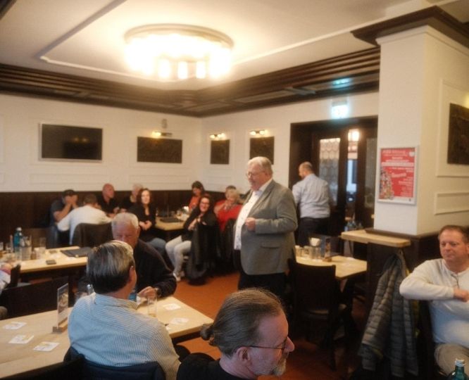 Ein älterer Mann im grauen Anzug steht in einem gut besuchten Restaurant und spricht zu einer Gruppe von Menschen, die an mehreren Tischen sitzen und aufmerksam zuhören. Die Atmosphäre wirkt gesellig und freundlich.