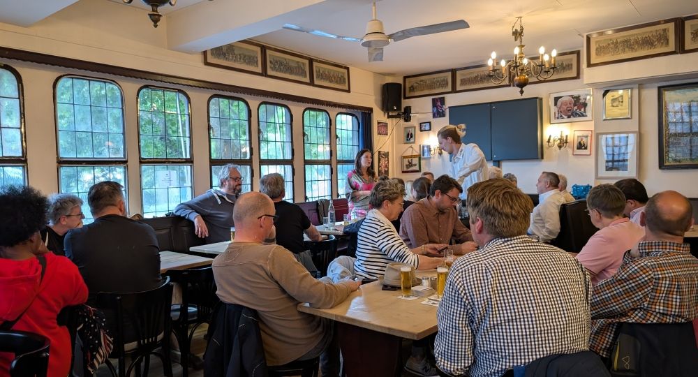 Eine Gruppe von Menschen sitzt in einem gemütlichen Restaurant mit großen Fenstern und Holztischen. Einige unterhalten sich, während ein Mann in weißem Hemd steht und spricht. Die Stimmung wirkt lebhaft und freundlich.