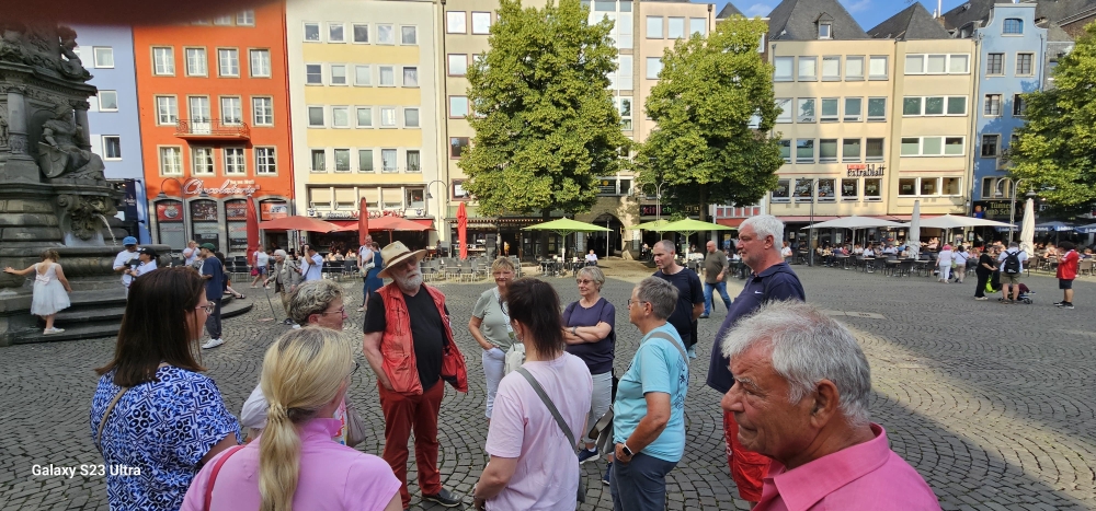 Eine Gruppe von Menschen steht auf einem Platz mit Kopfsteinpflaster in einer Altstadt. Im Hintergrund sind bunte Häuserfassaden, Straßencafés und Bäume zu sehen. Ein Mann mit Strohhut und roter Weste spricht mit der Gruppe.