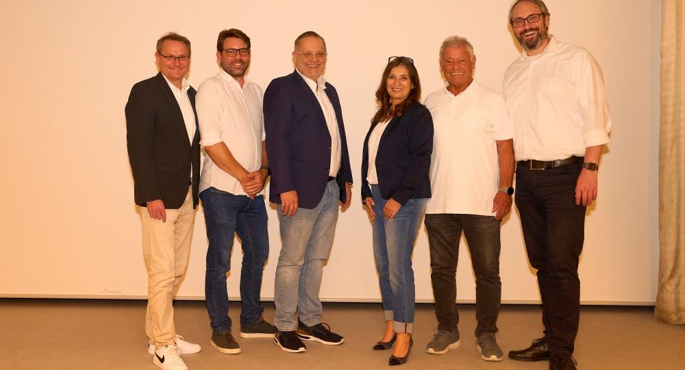 Sechs Personen stehen nebeneinander auf einer Bühne und lächeln in die Kamera, alle sind leger in Hemd, Jeans oder Sakko gekleidet.