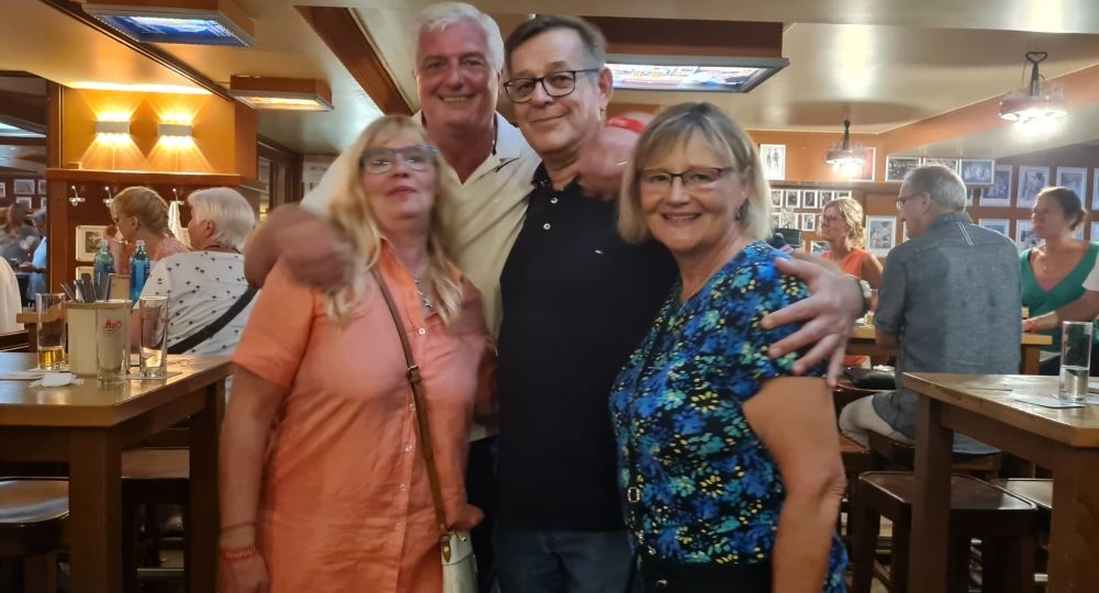 Zwei Männer und zwei Frauen stehen eng umarmt in einer gemütlichen Kneipe und lächeln in die Kamera.