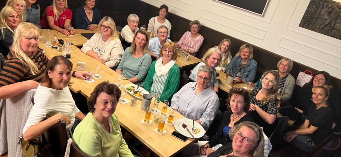 Eine große Gruppe von Frauen sitzt fröhlich an langen Tischen in einem Restaurant und schaut lächelnd in die Kamera.
