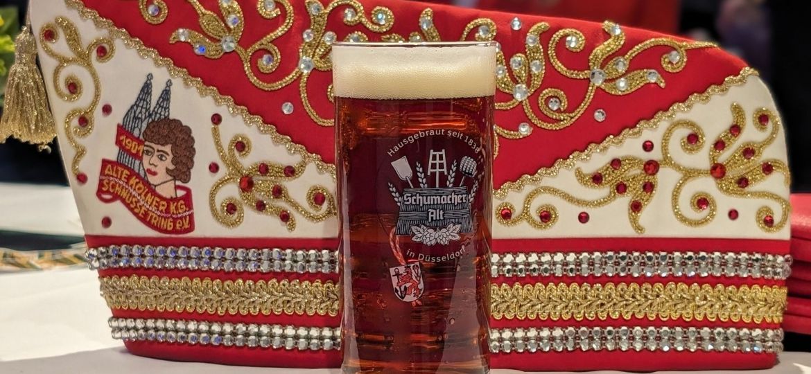 Ein Glas Altbier steht vor einer rot-weiß-gold bestickten Karnevalsmütze mit der Aufschrift „Alte Kölner K.G. Schnüsse Tring e.V.“.