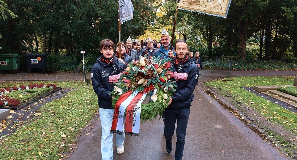 Zwei Männer tragen einen großen Kranz mit Schleifen, gefolgt von einer Gruppe mit Fahnen, bei einem Gedenkmarsch auf einem Friedhof.