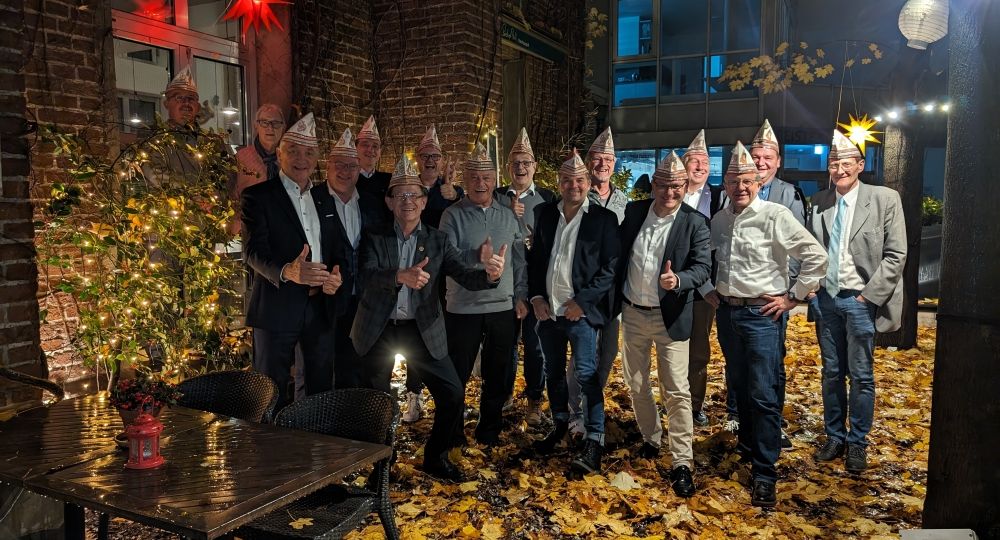Eine Gruppe von Männern mit Karnevalshüten steht lachend in einem herbstlich geschmückten Hof mit Lichterketten und bunten Sternen. Die schnüsse Tring weihnachtsfeier