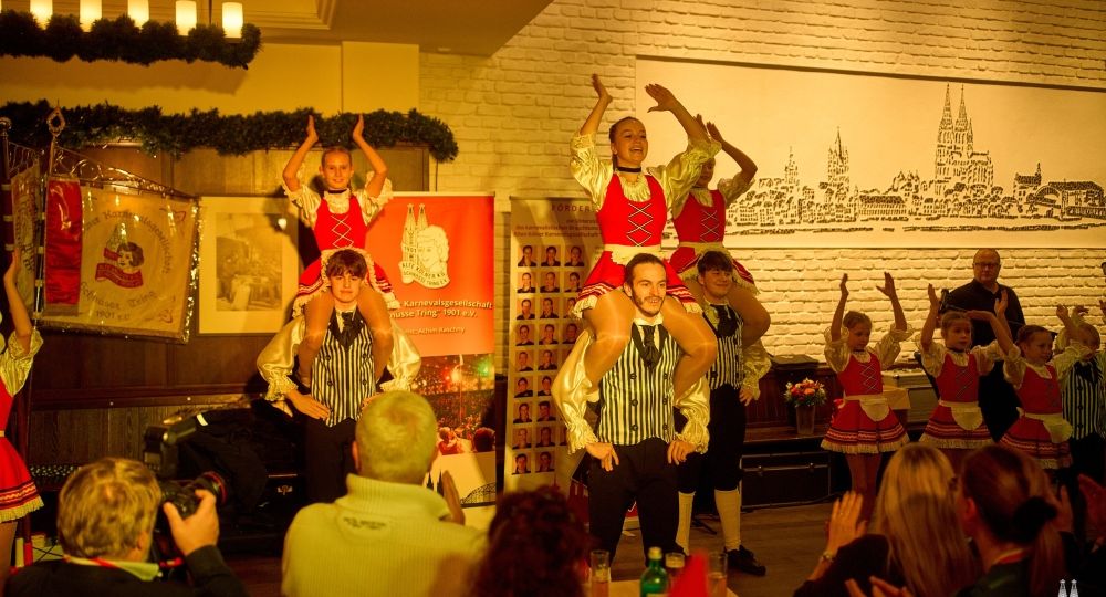 Eine Tanzgruppe in rot-weißen Kostümen führt auf einer Karnevalsveranstaltung eine Hebefigur vor, während das Publikum applaudiert.