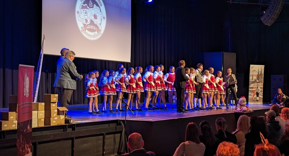 Eine Tanzgruppe in rot-weißen Kostümen steht auf einer Bühne bei einer Karnevalsveranstaltung, während das Publikum zuschaut.