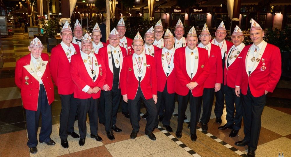 Eine Gruppe von Männern in roten Jacken und Karnevalshüten steht lächelnd in einer festlich beleuchteten Halle.
