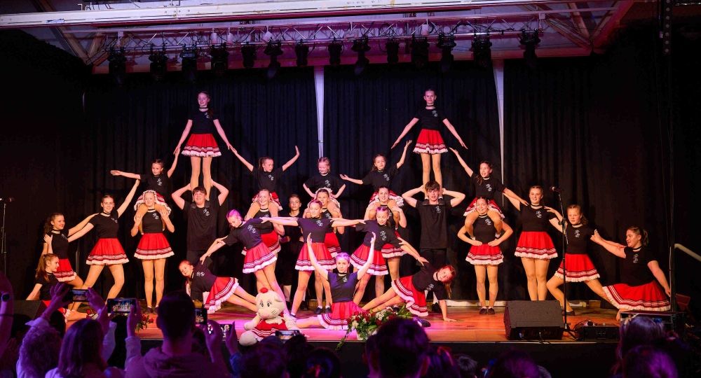 Eine große Tanzgruppe führt auf einer Bühne eine akrobatische Formation in rot-schwarzen Kostümen vor, während das Publikum applaudiert.