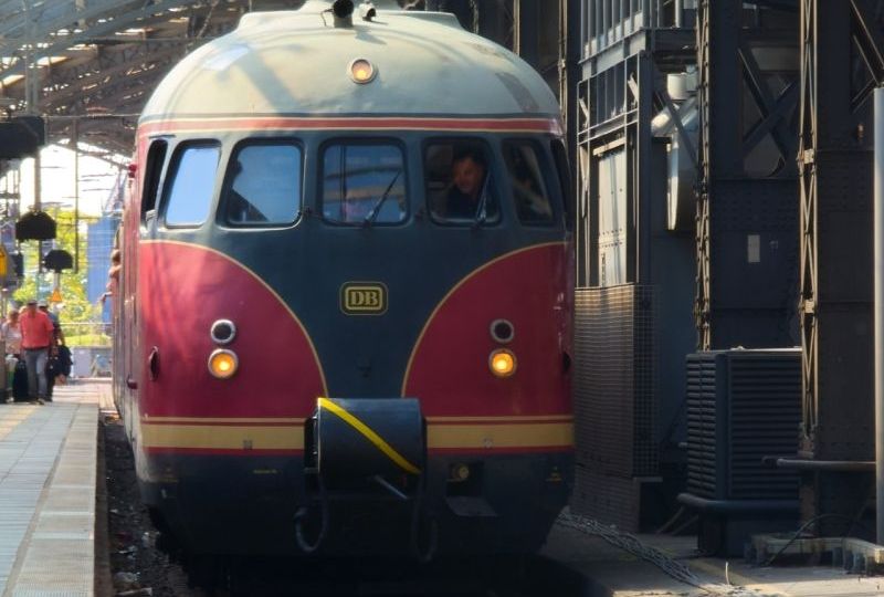 Eine rot-graue Lokomotive der Deutschen Bahn fährt in einen Bahnhof ein, während Menschen am Bahnsteig warten.