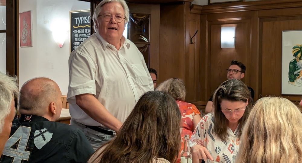 Ein Mann steht in einem Restaurant und spricht zu einer sitzenden Gruppe von Menschen, die gemeinsam essen.