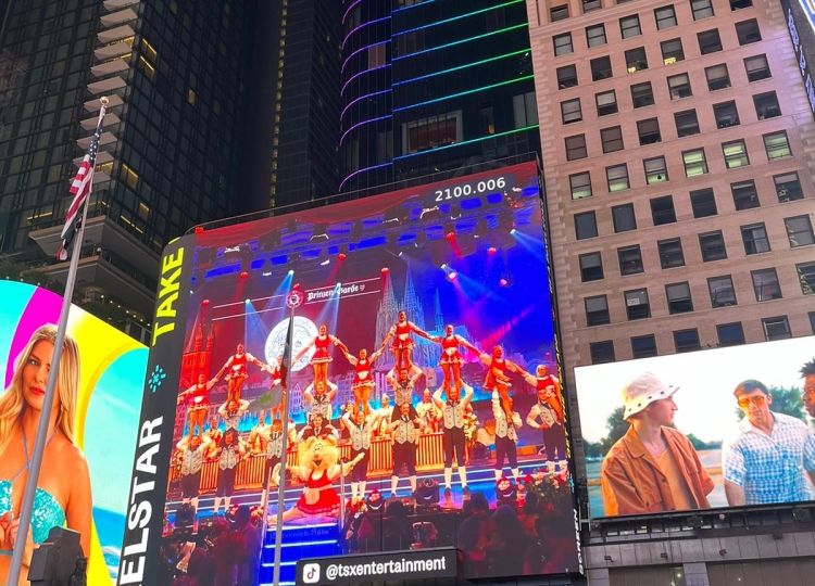 Eine große Menschenmenge steht am Times Square in New York, wo auf einem riesigen Bildschirm eine Tanzgruppe in roten Kostümen zu sehen ist.