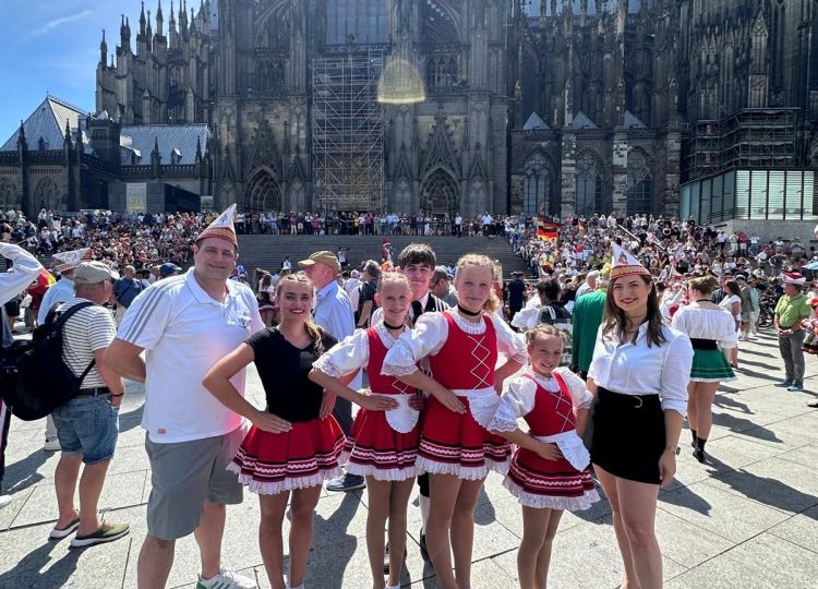 Eine Gruppe von Tänzerinnen in rot-weißen Kostümen und zwei Begleitpersonen posieren lächelnd vor dem Kölner Dom bei sonnigem Wetter.
