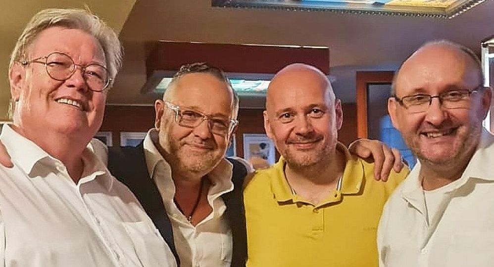 Vier Männer stehen dicht beieinander, lächeln in die Kamera und posieren in einer gemütlich beleuchteten Bar.