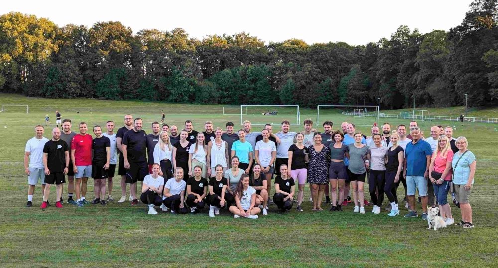 Eine große Gruppe von etwa 50 Personen steht gemeinsam auf einem Sportplatz im Freien. Im Hintergrund sind mehrere Fußballtore und Bäume zu sehen. Die Gruppe wirkt fröhlich und entspannt, einige Personen knien oder sitzen vorne, während andere stehen.