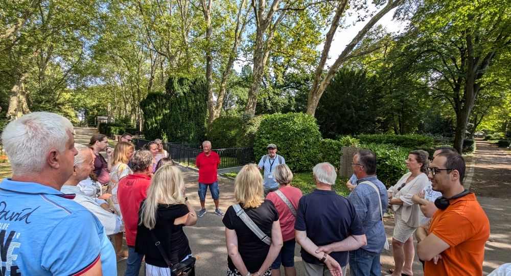 Eine Gruppe von Menschen steht in einem Park und hört einem Führer zu, der vor ihnen spricht.
