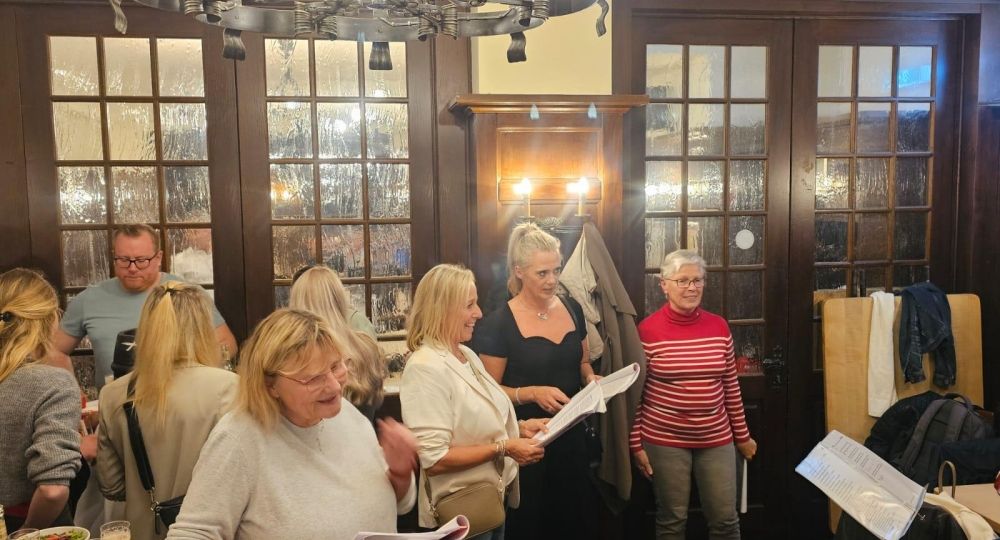 Mehrere Personen singen gemeinsam in einem gemütlichen Raum mit Holzverkleidung. Einige halten Liedblätter in der Hand, während auf den Tischen Gläser mit Bier stehen – die Szene wirkt gesellig und fröhlich.