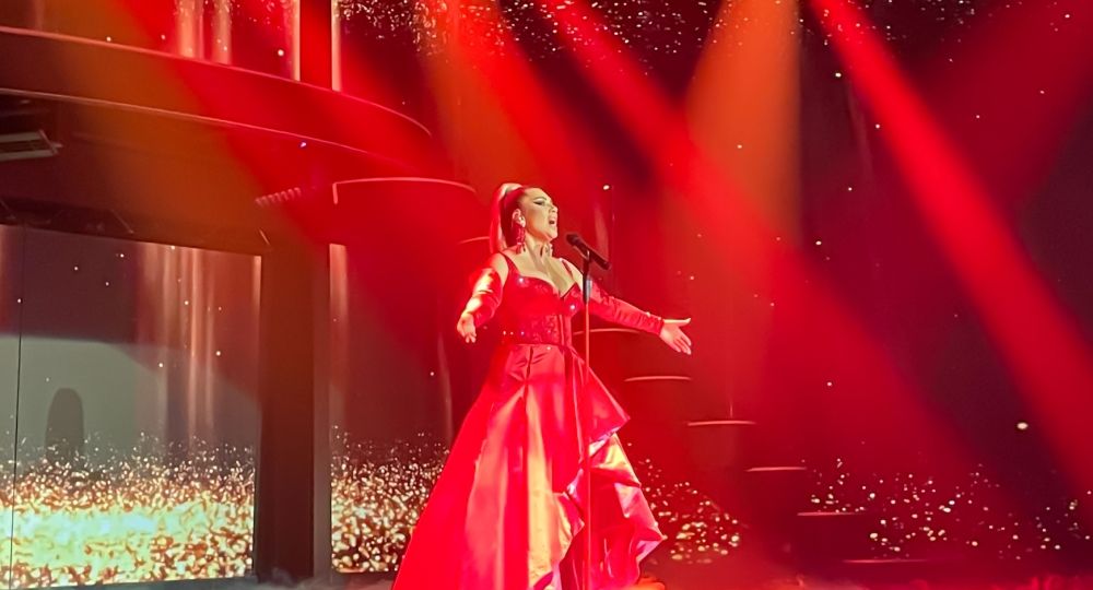 ChatGPT: Eine Sängerin steht auf der Bühne in eindrucksvollem roten Licht und trägt ein elegantes rotes Kleid – ein stimmungsvoller Moment voller Ausdruck und Emotion.