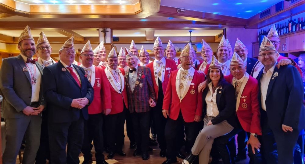 Das Bild zeigt eine große, fröhliche Gruppe von Menschen in festlicher Karnevalskleidung. Viele tragen rote Jacken, weiße Schärpen und traditionelle karnevalistische Mützen mit goldenen Verzierungen. Die Stimmung ist ausgelassen und feierlich – es scheint sich um ein offizielles Treffen oder eine Sitzung eines Karnevalsvereins zu handeln.